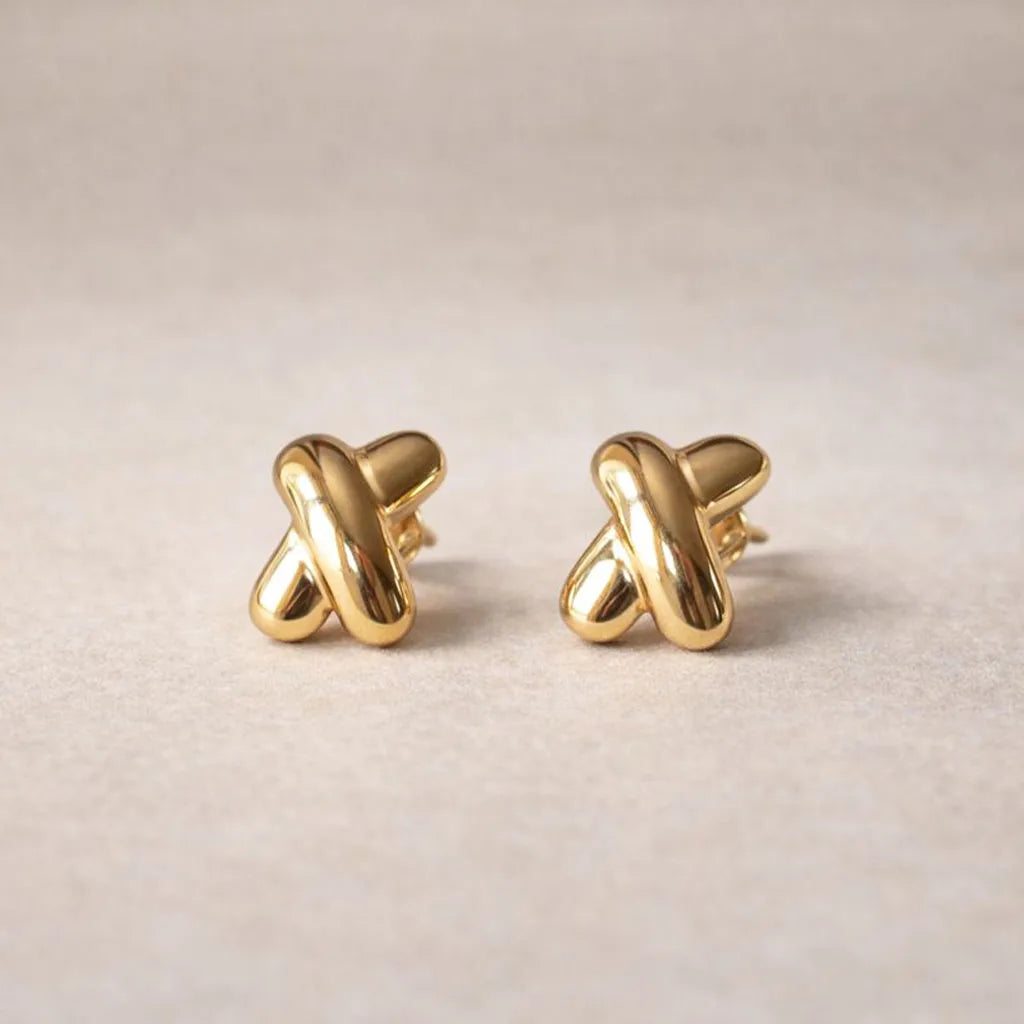 Boucles d'oreilles "Révèle toi", au design croix en acier inoxydable doré, photographiées sur un fond beige texturé.