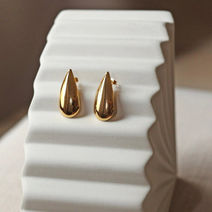 Boucles d'oreilles Larmes assumées en  acier doré, en forme de goutte, posées sur un support beige texturé.