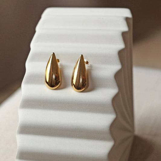 Boucles d'oreilles Larmes assumées en  acier doré, en forme de goutte, posées sur un support beige texturé.