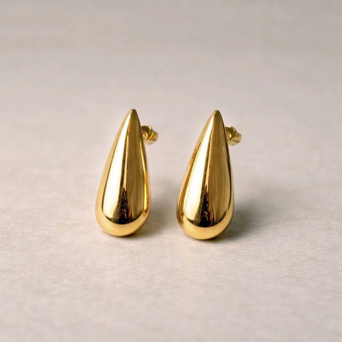 Boucles d'oreilles Larmes assumées en  acier doré, en forme de goutte, posées sur un fond  beige texturé.