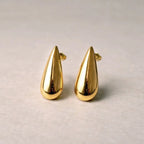 Boucles d'oreilles Pure en  acier doré, en forme de goutte, posées sur un fond  beige texturé.
