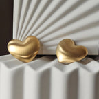 Zoom sur les boucles d'oreilles "S'aimer", en maxi cœurs dorés mat, en acier Inoxydable, posées sur un fond beige texturé.