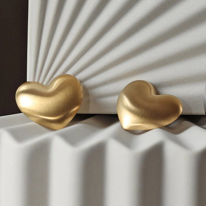 Zoom sur les boucles d'oreilles "S'aimer", en maxi cœurs dorés mat, en acier Inoxydable, posées sur un fond beige texturé.