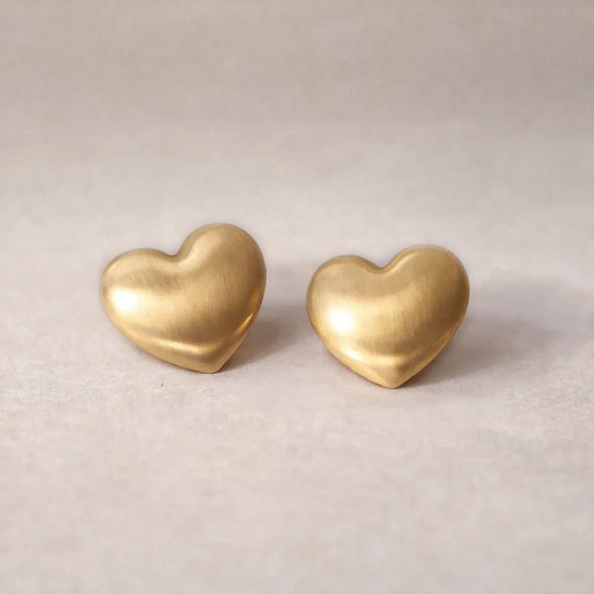 Zoom sur les boucles d'oreilles "S'aimer", en maxi cœurs dorés mat, en acier Inoxydable, posées sur un fond beige texturé.