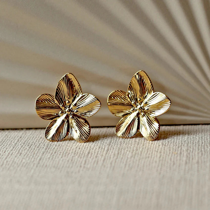 Boucles d'oreilles Fleur de peau en acier inoxydable dorée, motif floral oversize, posées sur un tissu beige texturé.
