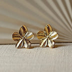 Boucles d'oreilles Fleur de peau en acier inoxydable dorée, motif floral oversize, posées sur un tissu beige texturé.