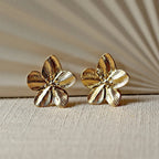Boucles d'oreilles Fleur de peau en acier inoxydable dorée, motif floral oversize, posées sur un tissu beige texturé.