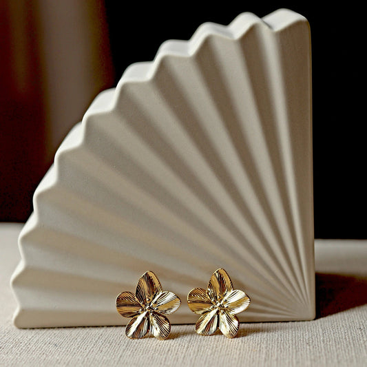 Boucles d'oreilles Fleur de peau en acier inoxydable dorée, grand motif floral, posées sur un tissu beige texturé.