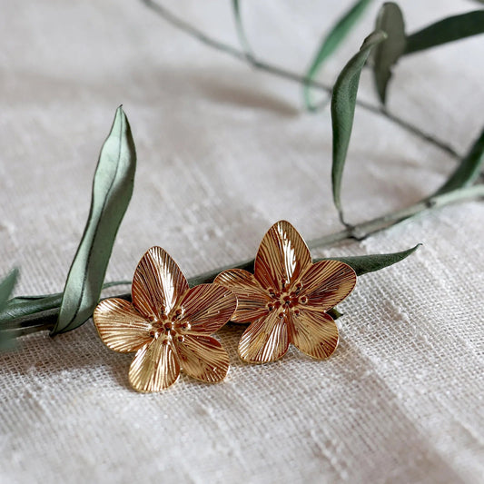 Boucles d'oreilles Fleur de peau en acier inoxydable dorée, motif floral oversize, posées sur une branche de feuille et un tissu beige texturé.