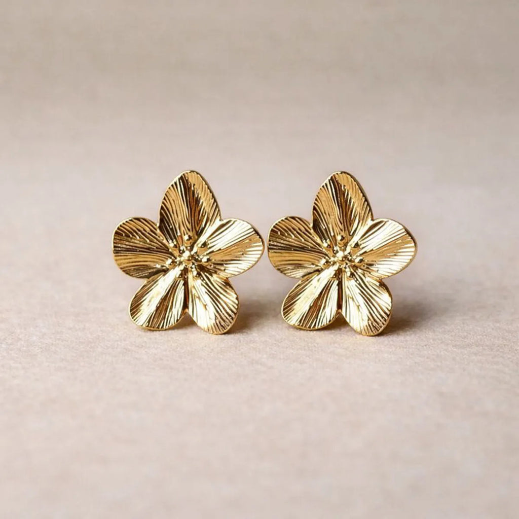 Boucles d'oreilles Fleurie en acier inoxydable dorée, motif floral oversize, posées sur un tissu beige texturé.