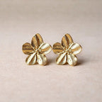 Boucles d'oreilles Fleurie en acier inoxydable dorée, motif floral oversize, posées sur un tissu beige texturé.