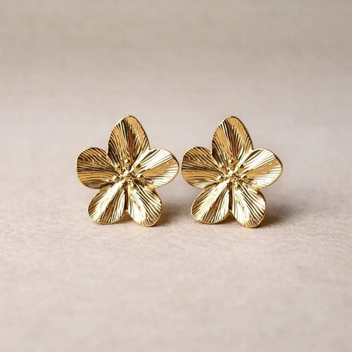 Boucles d'oreilles Fleurie en acier inoxydable dorée, motif floral oversize, posées sur un tissu beige texturé.