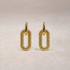 Boucles d'oreilles "Modulable" en acier doré, au design modulable, photographiées sur un tissu beige texturé.