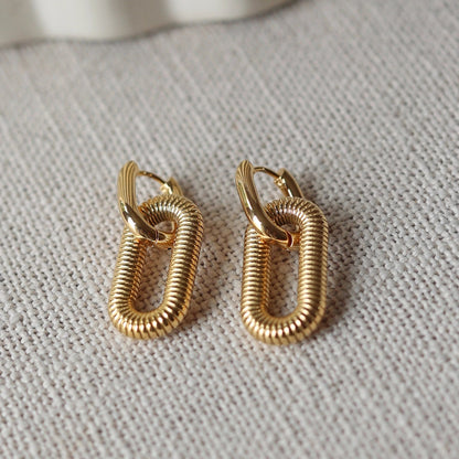 Boucles d'oreilles "J'existe" en acier doré, au design modulable, photographiées sur un tissu beige texturé.