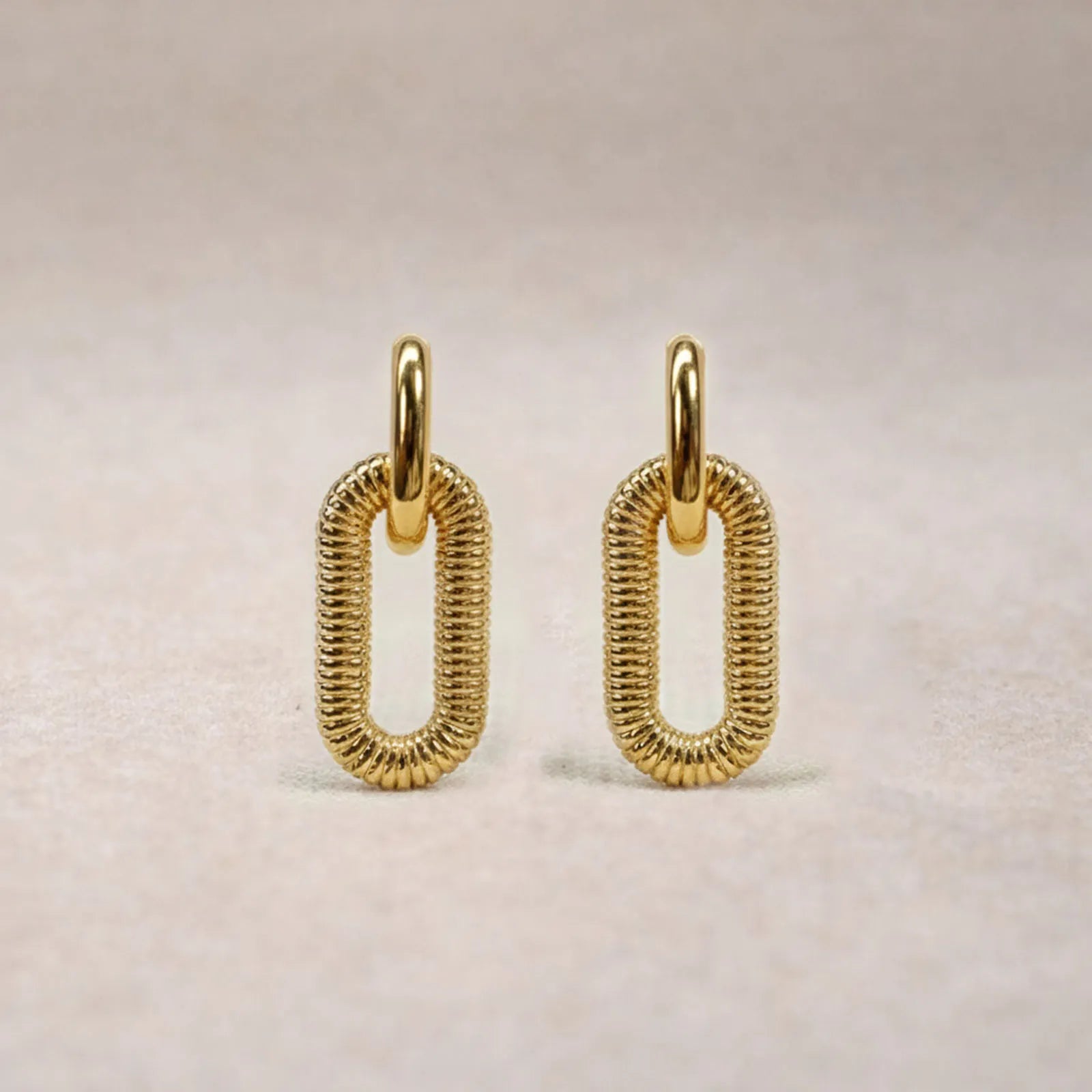 Boucles d'oreilles "J'existe" en acier doré, au design modulable, photographiées sur un tissu beige texturé.