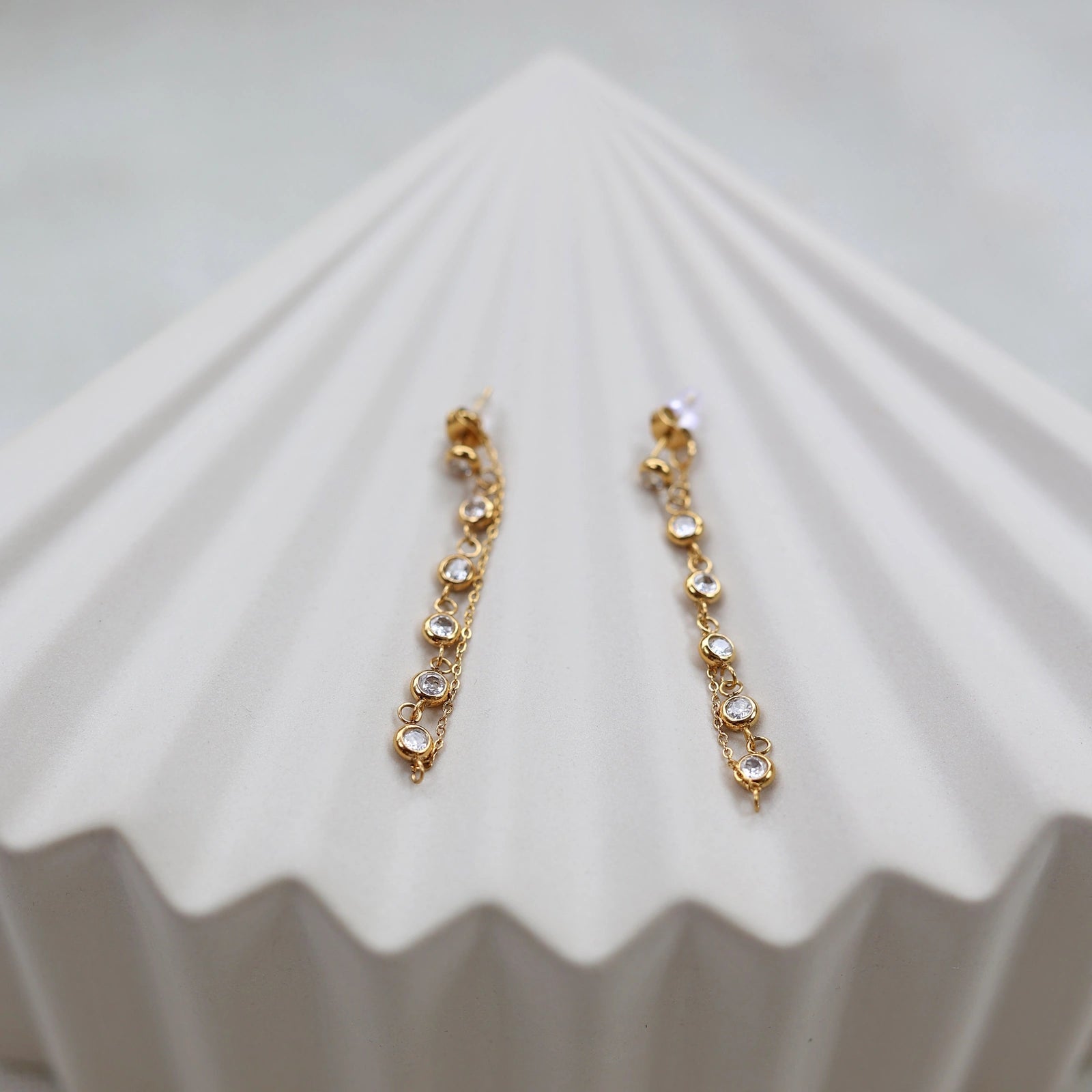 Boucles d'oreilles dorées Eclat, pendantes, serties de strass brillants, en acier. Photographiées sur un support beige texturé.