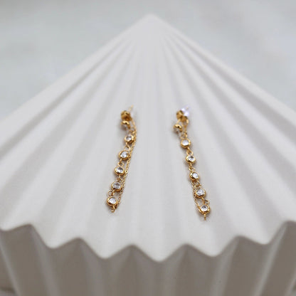 Boucles d'oreilles dorées Eclat, pendantes, serties de strass brillants, en acier. Photographiées sur un support beige texturé.