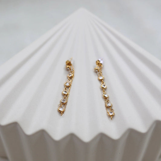 Boucles d'oreilles dorées Eclat, pendantes, serties de strass brillants, en acier. Photographiées sur un support beige texturé.