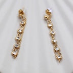 Boucles d'oreilles dorées Eclat, pendantes et serties de strass brillants, en acier. Photographiées en gros plan sur un support beige texturé.