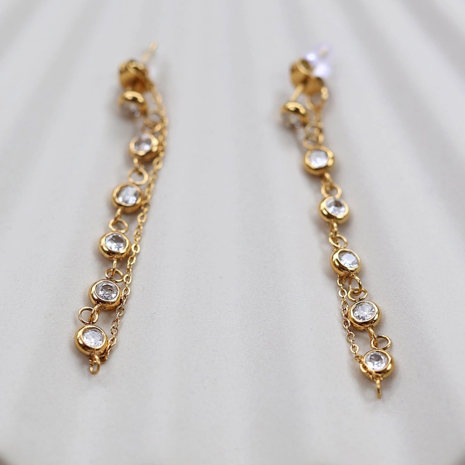 Boucles d'oreilles dorées Eclat, pendantes et serties de strass brillants, en acier. Photographiées en gros plan sur un support beige texturé.