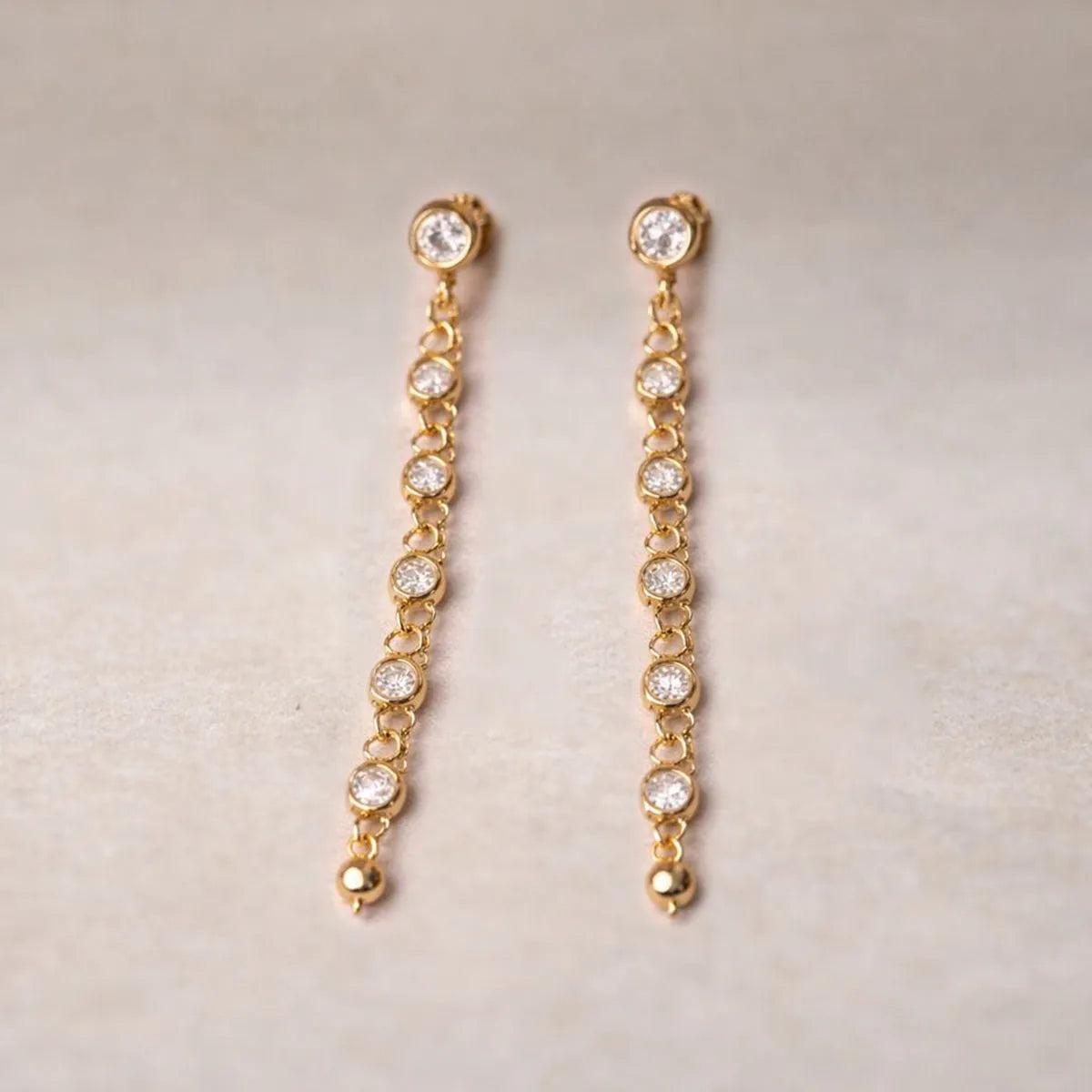 Boucles d'oreilles dorées Eclat, pendantes, serties de strass brillants, en acier. Photographiées sur un fons beige texturé.