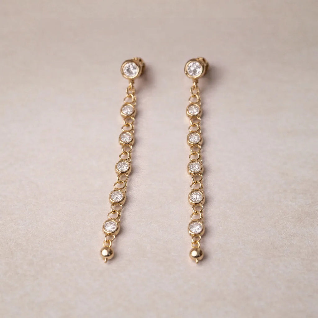 Boucles d'oreilles dorées Rivière, pendantes, serties de strass brillants, en acier. Photographiées sur un fons beige texturé.