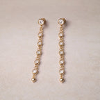 Boucles d'oreilles dorées Rivière, pendantes, serties de strass brillants, en acier. Photographiées sur un fons beige texturé.