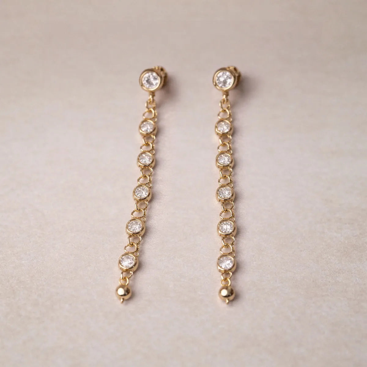 Boucles d'oreilles dorées Rivière, pendantes, serties de strass brillants, en acier. Photographiées sur un fons beige texturé.