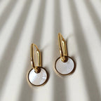 Zoom sur les boucles d'oreilles "Solstice", en acier doré, pendentifs cercles personnalisables à effet nacré, posées sur un fond beige texturé.