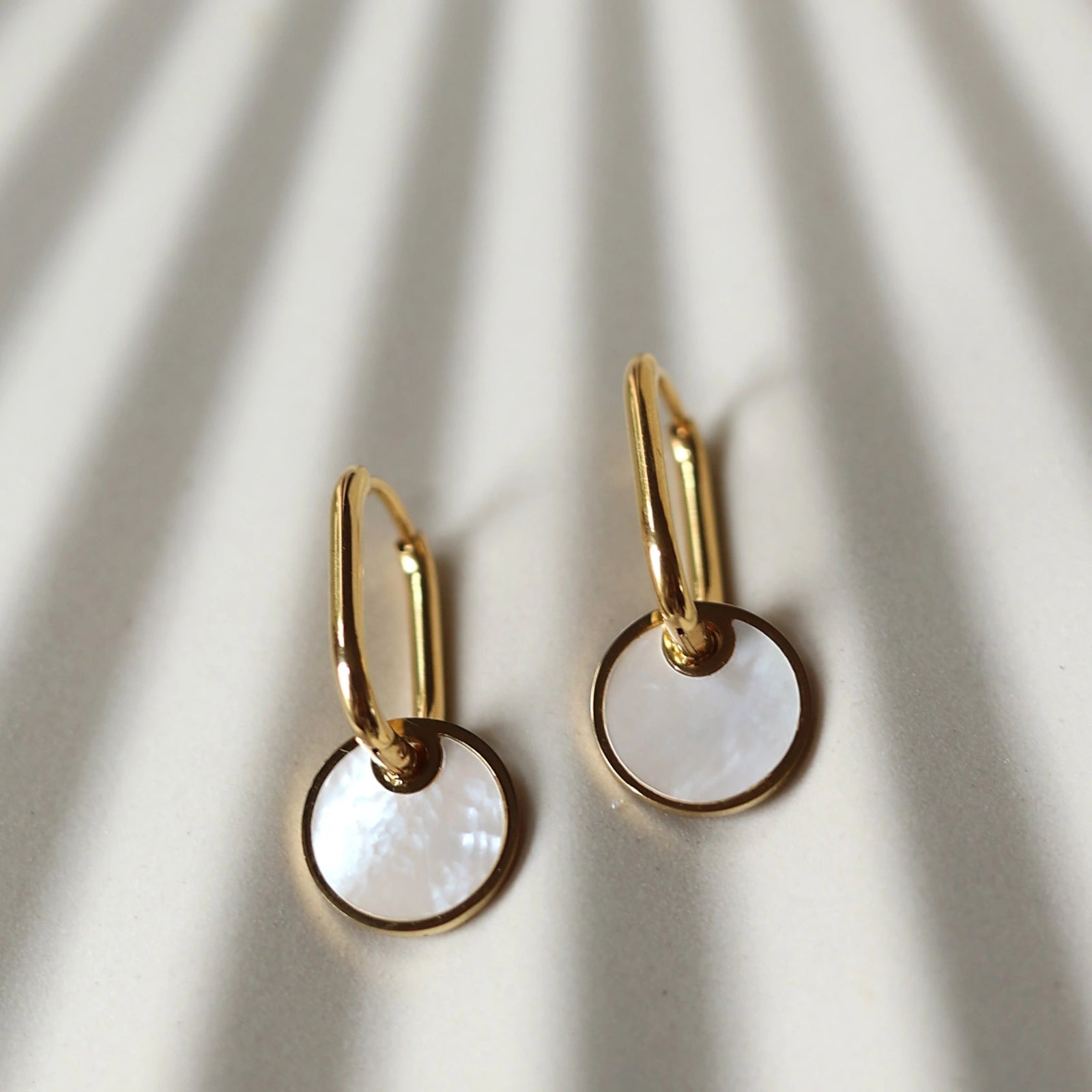 Zoom sur les boucles d'oreilles "Solstice", en acier doré, pendentifs cercles personnalisables à effet nacré, posées sur un fond beige texturé.