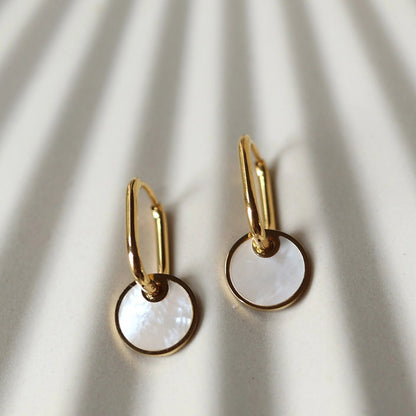Zoom sur les boucles d'oreilles "Solstice", en acier doré, pendentifs cercles personnalisables à effet nacré, posées sur un fond beige texturé.