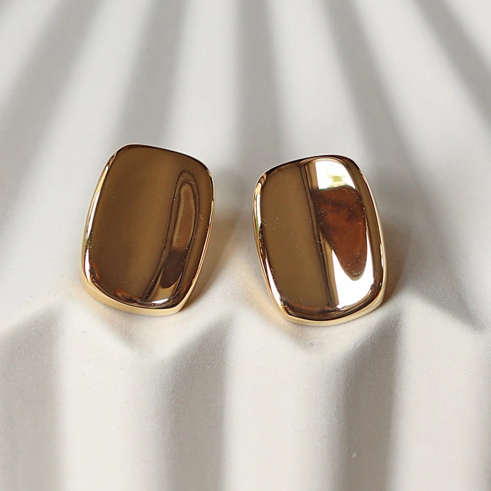Vue rapprochée des boucles d’oreilles Fierté minimalistes rectangulaires  en acier inoxydable doré, posées à plat sur un support beige texturé.