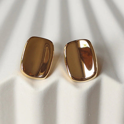 Vue rapprochée des boucles d’oreilles Fierté minimalistes rectangulaires  en acier inoxydable doré, posées à plat sur un support beige texturé.