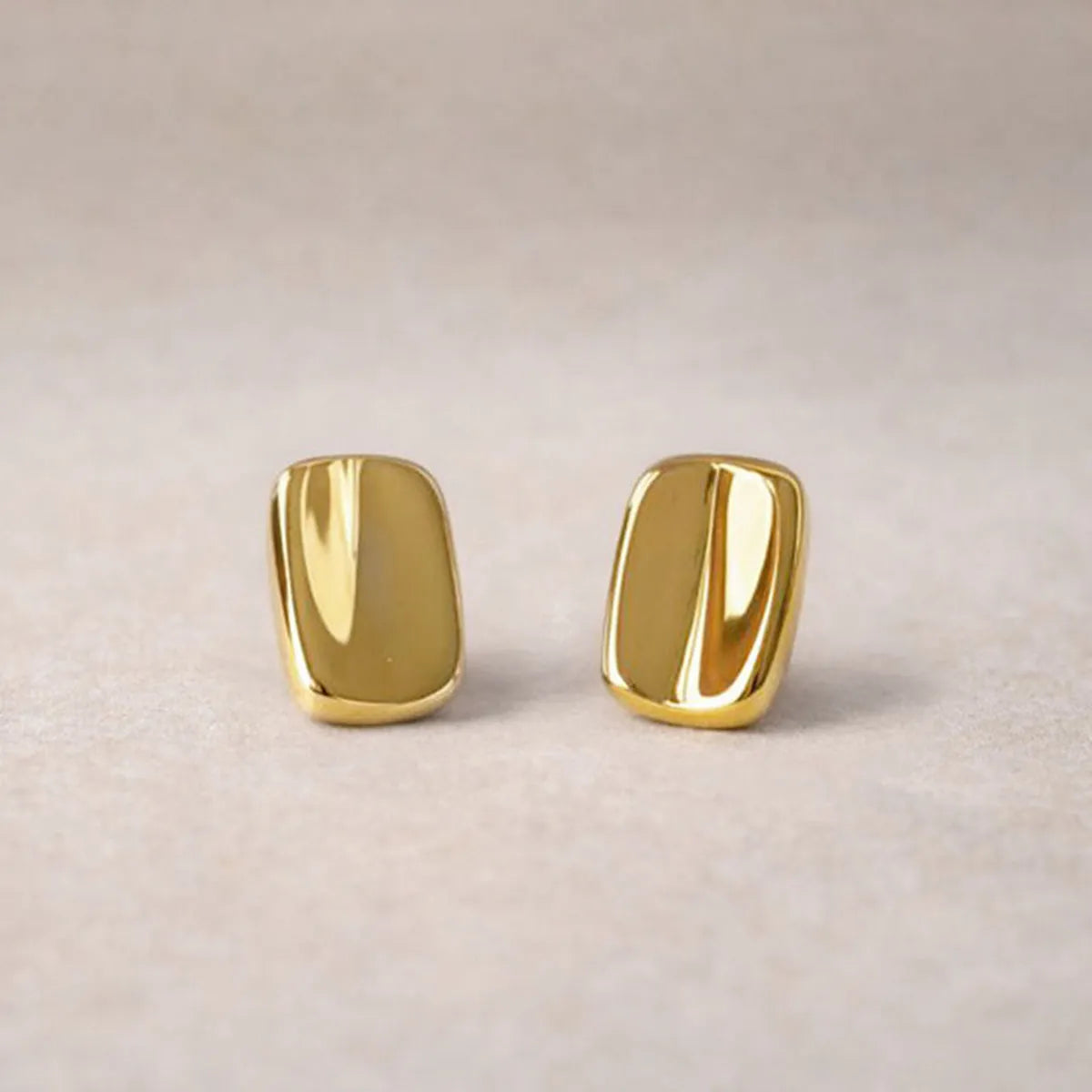 Vue rapprochée des boucles d’oreilles Fierté minimalistes rectangulaires  en acier inoxydable doré, posées sur un fond beige texturé.
