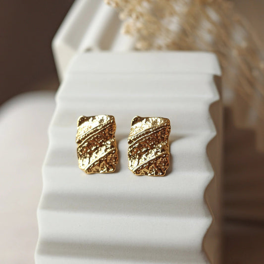 Boucles d'oreilles Affirmation rectangulaires texturées en acier doré, photographiées sur un fond beige texturé.