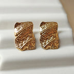 Zoom sur les boucles d'oreilles Affirmation, rectangulaires et texturées en acier doré, posées sur un fond beige texturé.