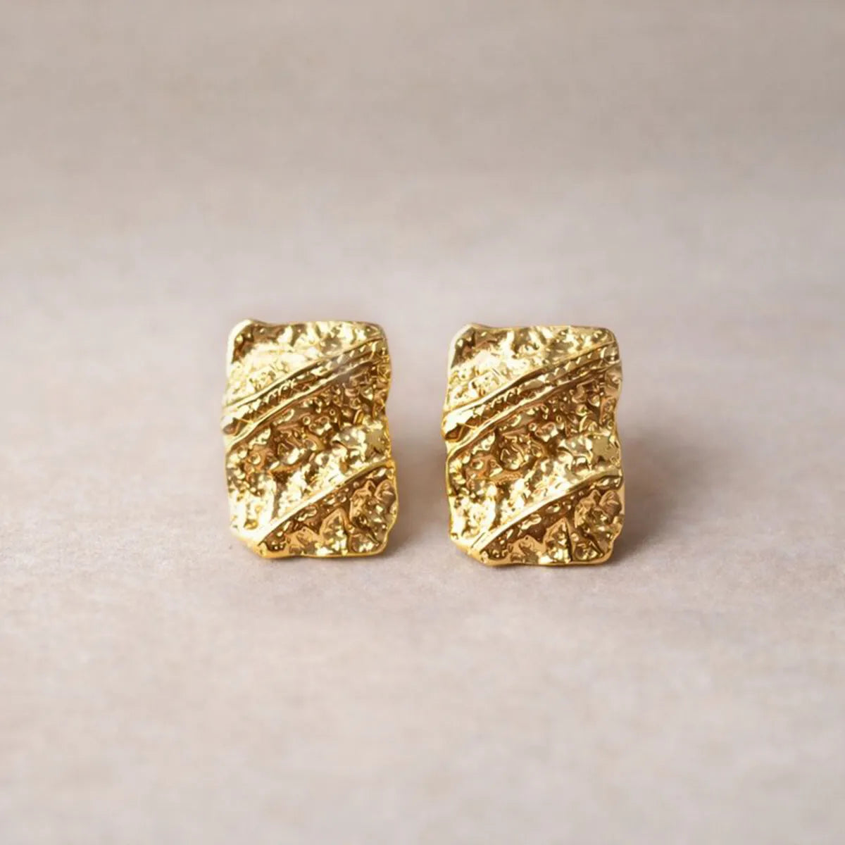 Boucles d'oreilles Affirmation rectangulaires texturées en acier doré, photographiées sur un fond beige texturé.