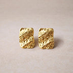 Boucles d'oreilles Relief rectangulaires texturées en acier doré, photographiées sur un fond beige texturé.
