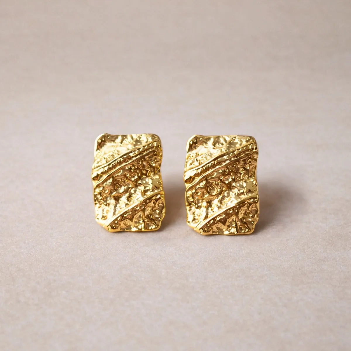 Boucles d'oreilles Relief rectangulaires texturées en acier doré, photographiées sur un fond beige texturé.