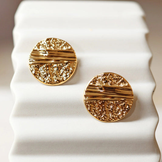 Boucles d'oreilles Rayonnante, en acier inoxydable doré, au design rond texturé et éclatant. Gros plan sur un support beige texturé.