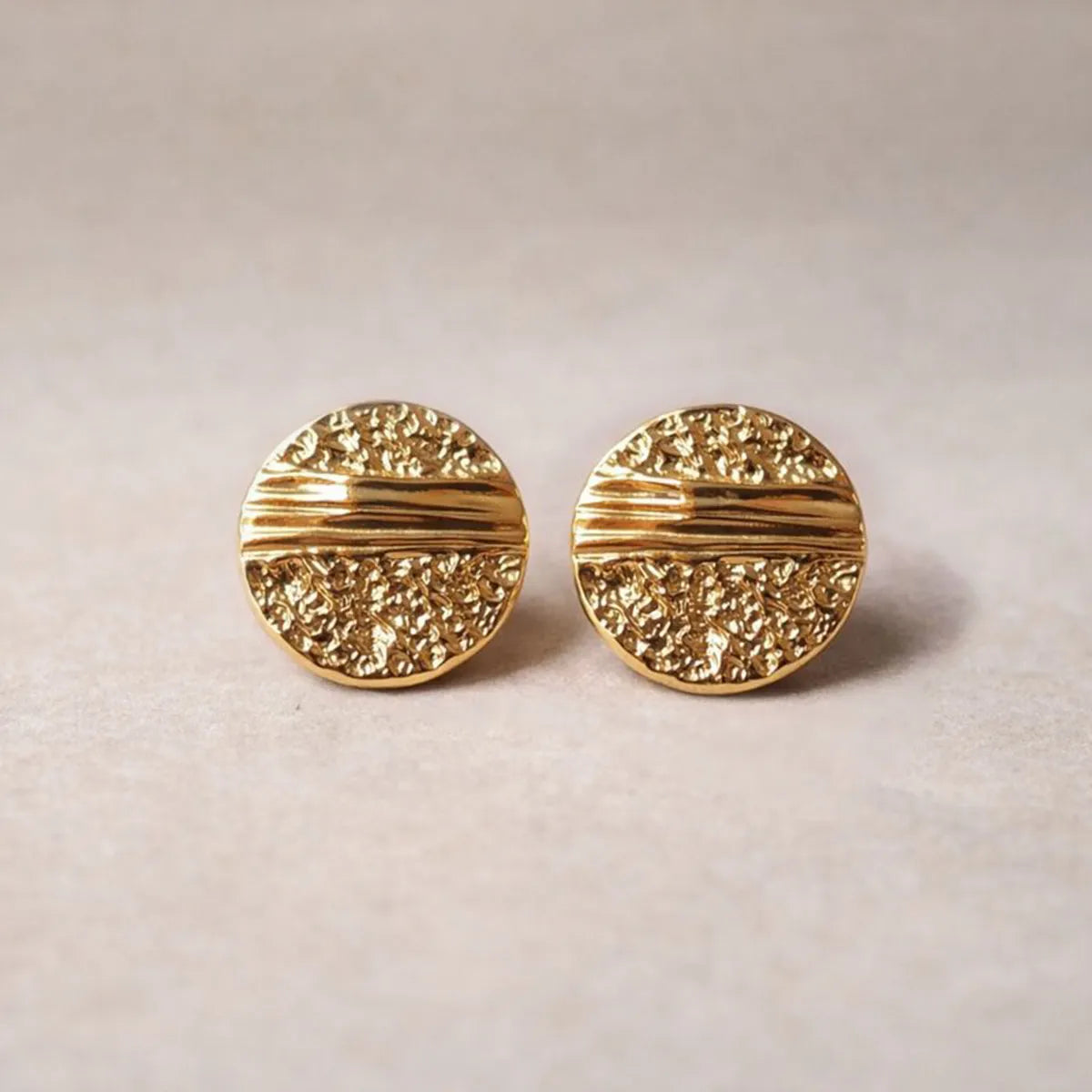 Boucles d'oreilles Rayonnante, en acier inoxydable doré, au design rond texturé et éclatant. Gros plan sur un fons beige texturé.