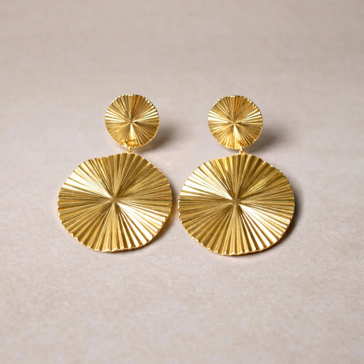 Vue d'ensemble des boucles d'oreilles Rayonnante pendantes au design solaire strié en acier inoxydable doré, posées à plat sur un fond en toile beige texturé.