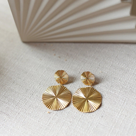 Vue d'ensemble des boucles d'oreilles Vivante pendantes au design solaire strié en acier inoxydable doré, posées à plat sur un fond en toile beige texturé.