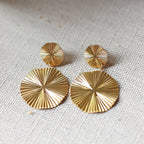 Vue rapprochée des boucles d'oreilles Vivante pendantes au design solaire strié en acier inoxydable doré, posées à plat sur un fond beige texturé.