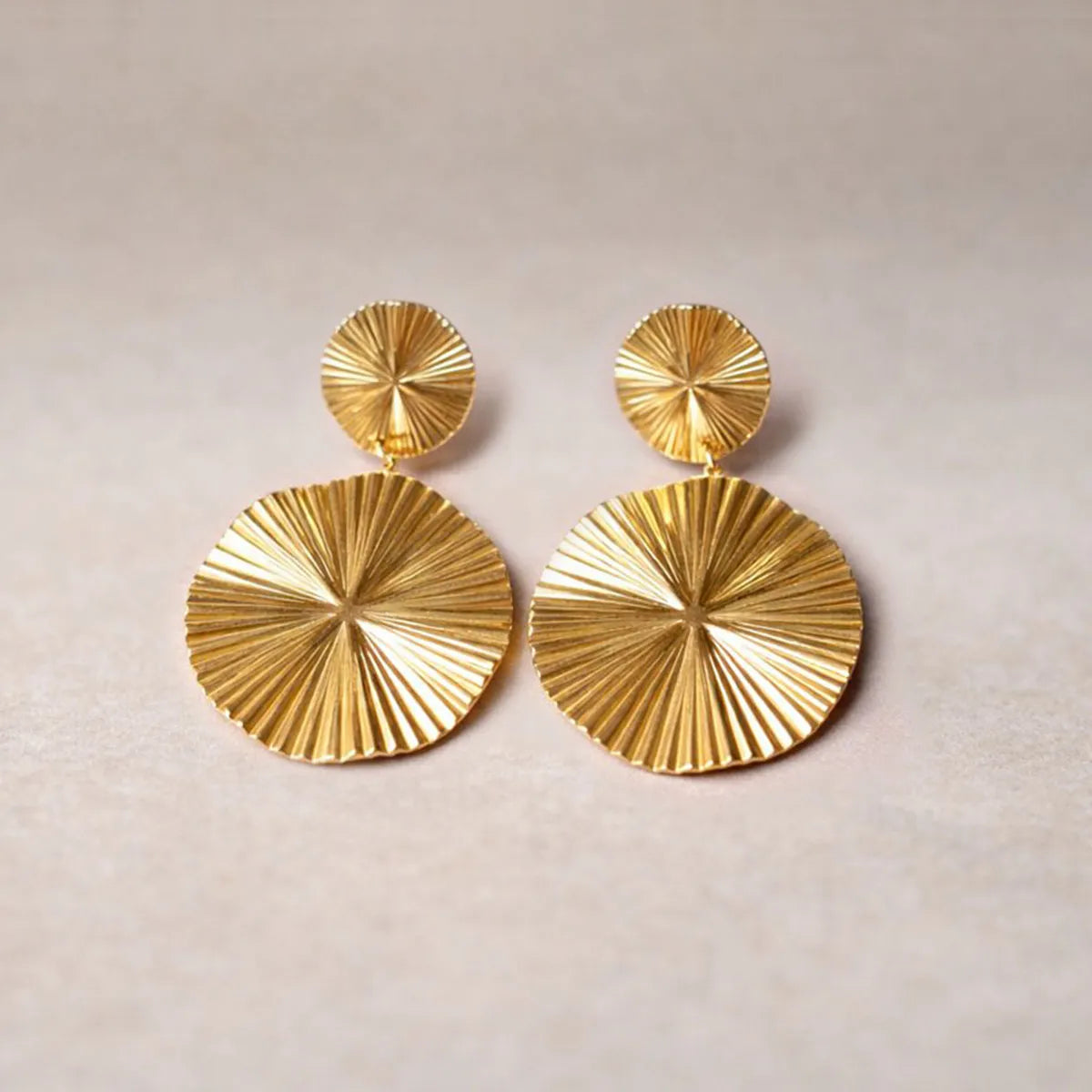 Vue d'ensemble des boucles d'oreilles Vivante pendantes au design solaire strié en acier inoxydable doré, posées à plat sur un fond en toile beige texturé.