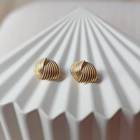 Vue rapprochée des boucles d’oreilles Métamorphose torsadées en acier inoxydable doré, posées à plat sur une structure beige texturée. 
