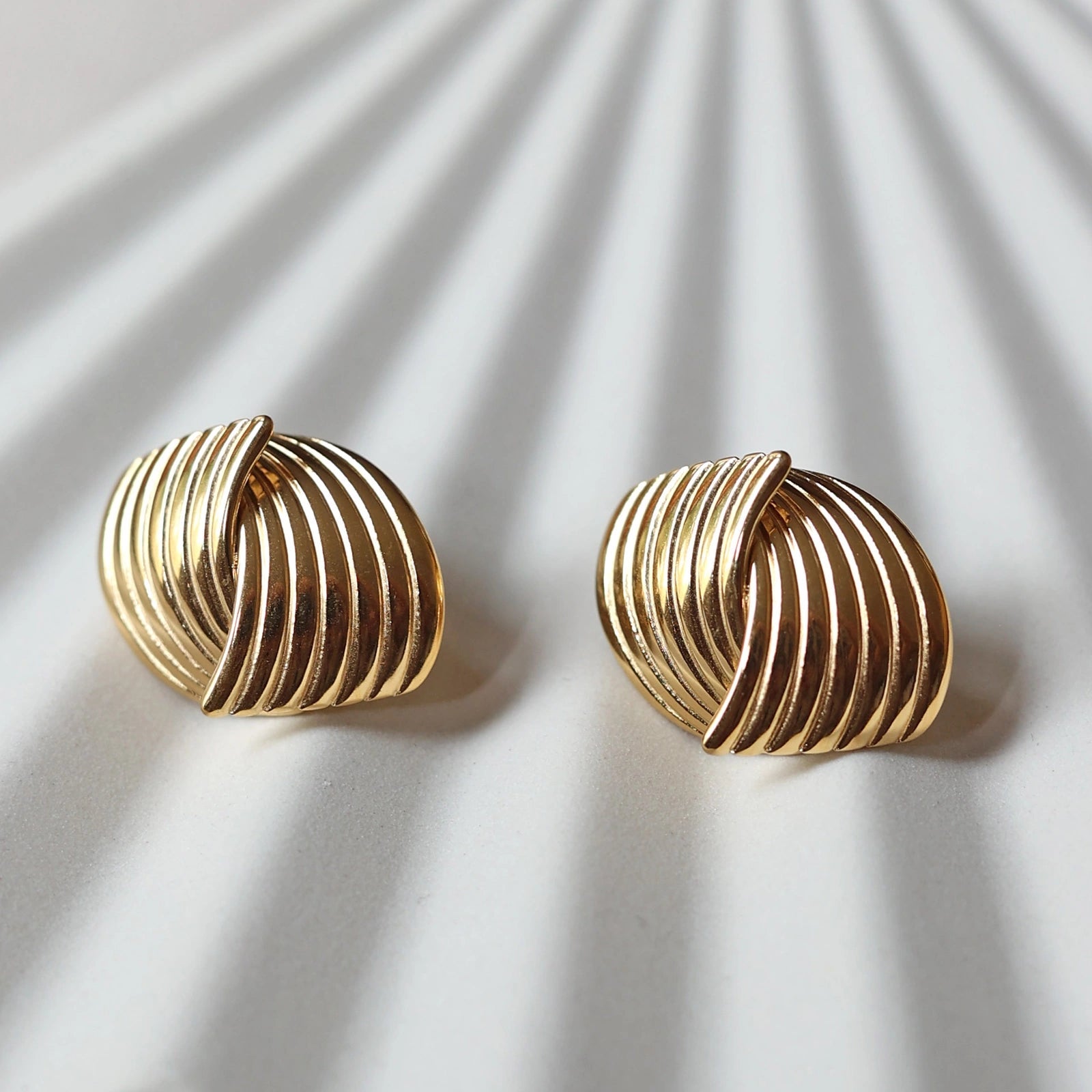 Vue rapprochée des boucles d’oreilles Metémorphose torsadées en acier inoxydable doré, posées à plat sur une structure beige texturée. 