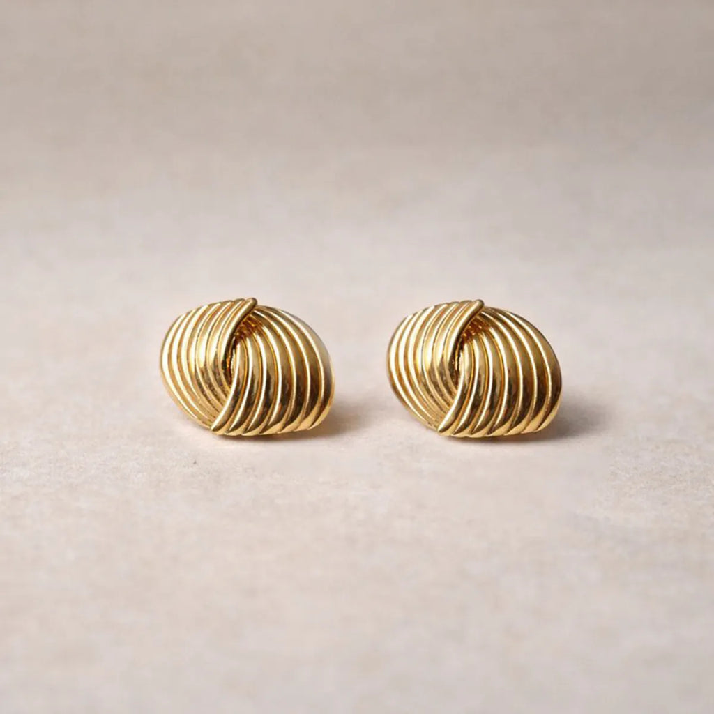 Vue rapprochée des boucles d’oreilles Metémorphose torsadées en acier inoxydable doré, posées sur un fond beige texturé. 