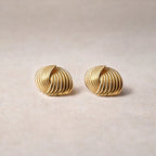 Vue rapprochée des boucles d’oreilles Metémorphose torsadées en acier inoxydable doré, posées sur un fond beige texturé. 