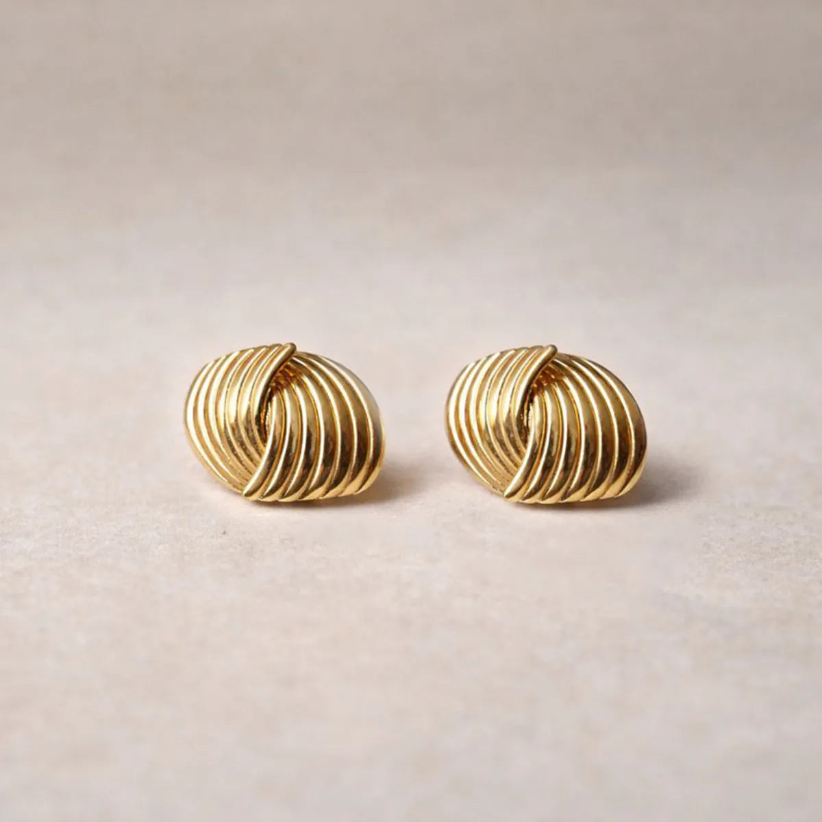 Vue rapprochée des boucles d’oreilles Metémorphose torsadées en acier inoxydable doré, posées sur un fond beige texturé. 